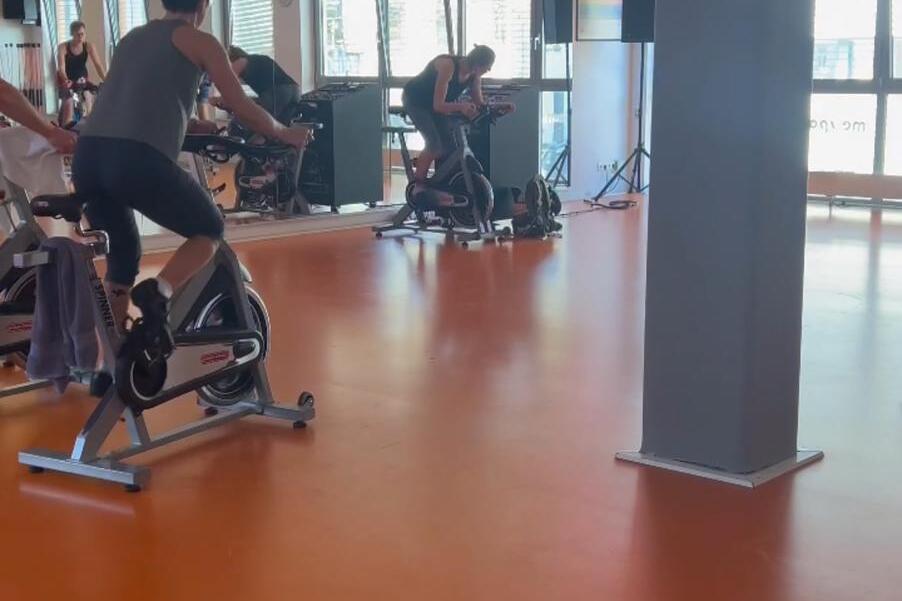 Menschen sitzen auf Fahrrädern in einem Fitnessraum mit orangefarbenem Boden und großen Fenstern.