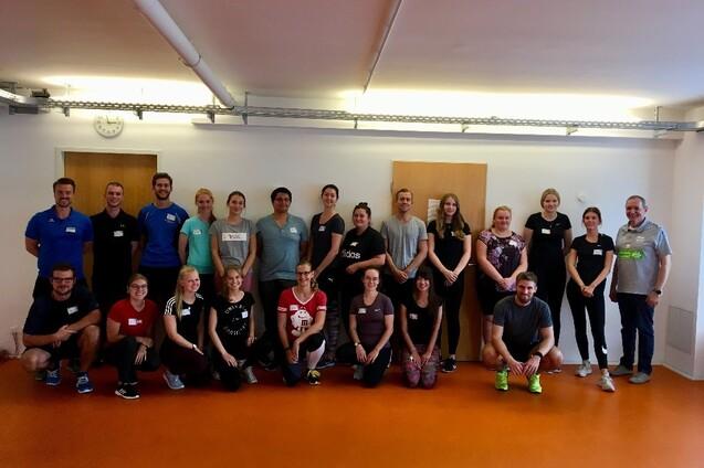 Gruppenfoto von 22 Personen in Sportkleidung, lächelnd, in einem hellen Raum mit orangefarbenem Boden.