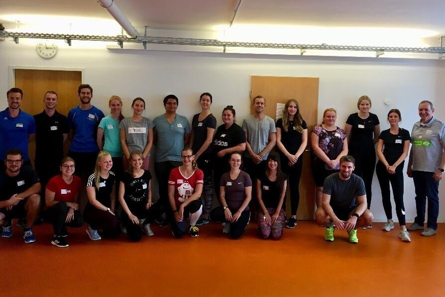 Gruppenfoto von 22 Personen in Sportkleidung, lächelnd, in einem hellen Raum mit orangefarbenem Boden.