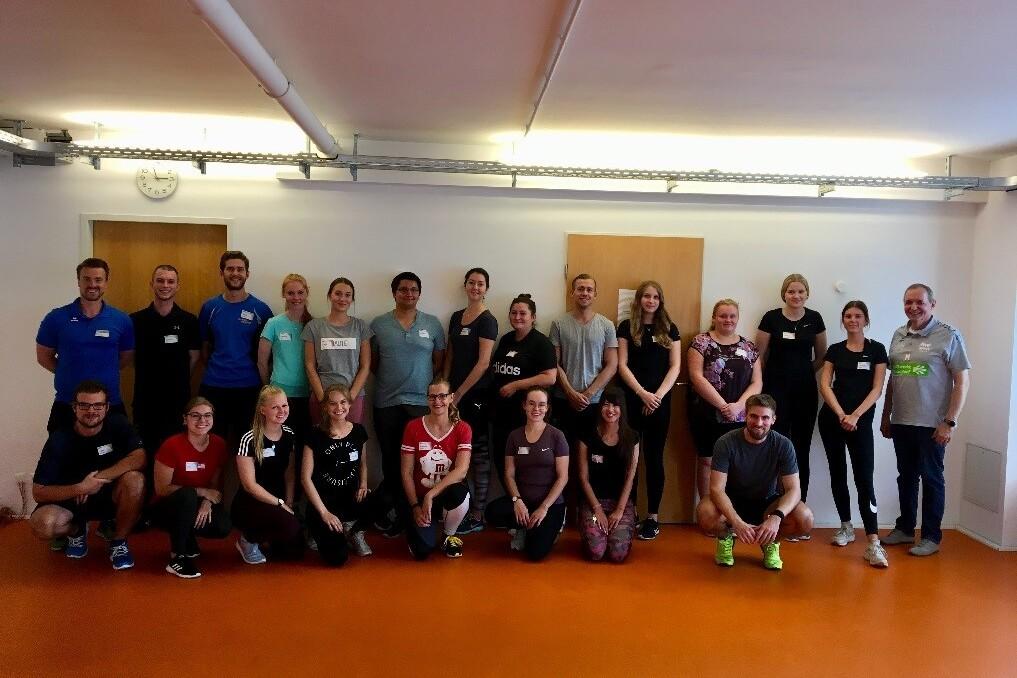 Gruppenfoto von 22 Personen in Sportkleidung, lächelnd, in einem hellen Raum mit orangefarbenem Boden.