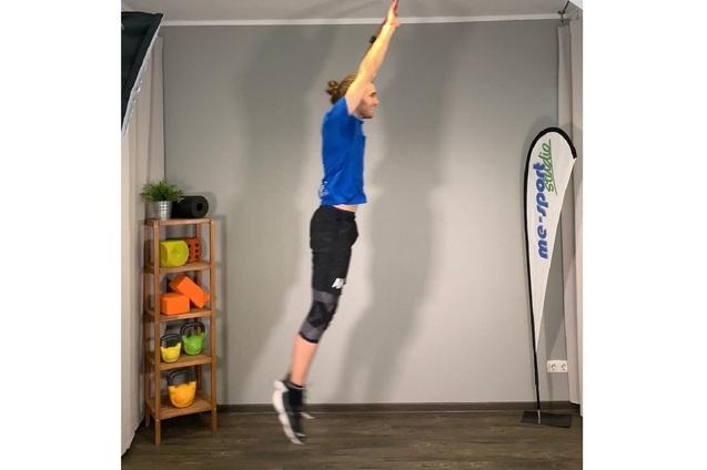 Mann springt in Sportkleidung mit erhobenen Armen in einem Fitnessstudio mit Trainingsgeräten im Hintergrund.