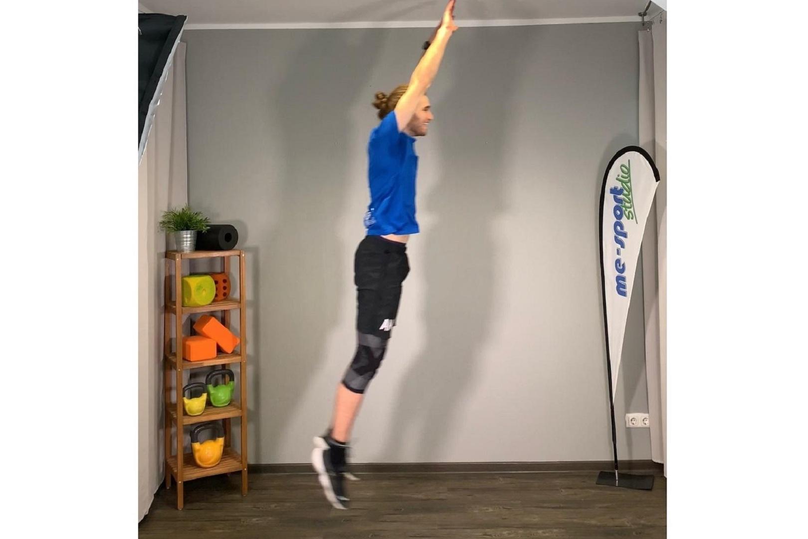Mann springt in Sportkleidung mit erhobenen Armen in einem Fitnessstudio mit Trainingsgeräten im Hintergrund.