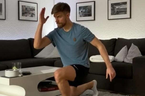 Junger Mann in Sportkleidung f&uuml;hrt eine Dehn&uuml;bung im Wohnzimmer auf einem Teppich aus, Couch und Bilder im Hintergrund.