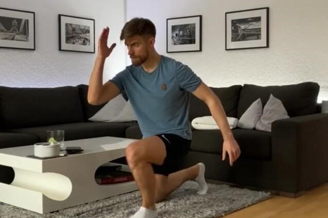Junger Mann in Sportkleidung führt eine Dehnübung im Wohnzimmer auf einem Teppich aus, Couch und Bilder im Hintergrund.