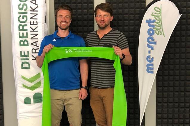 Zwei Männer halten ein grünes Band, stehen vor Flaggen der BKK und eines E-Sport-Studios in einem mit Schaumstoff ausgekleideten Raum.