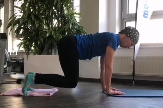 Frau in Sportkleidung führt eine Übungen auf einer blauen Matte in einem Fitnessraum mit großen Fenstern aus.