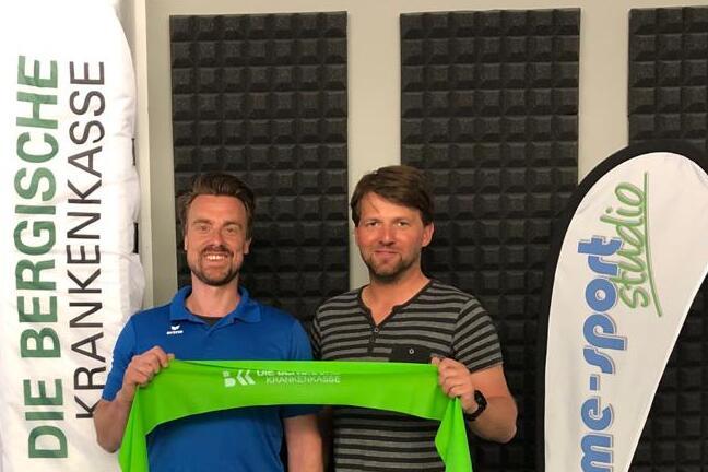 Zwei M&auml;nner halten ein gr&uuml;nes Band, stehen vor Flaggen der BKK und eines E-Sport-Studios in einem mit Schaumstoff ausgekleideten Raum.