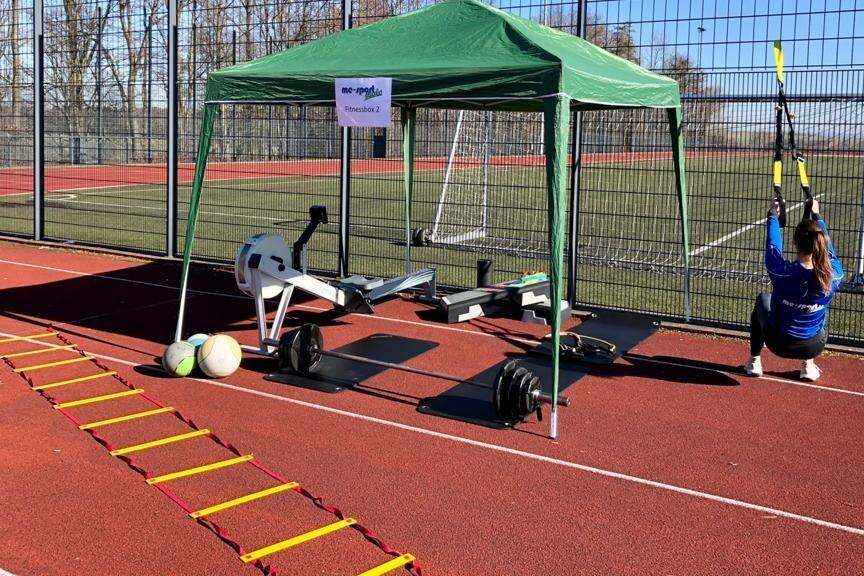 Sportbereich mit gr&uuml;nem Zelt, Fitnessger&auml;ten, einer Person mit TRX-B&auml;ndern und Trainingsmaterial auf rotem Platz.