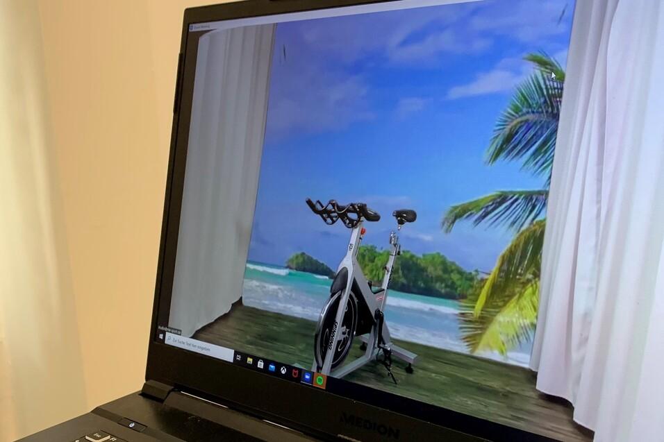 Virtuelles Fitnessgerät steht auf einem Tisch, im Hintergrund eine tropische Strandlandschaft mit Palmen.