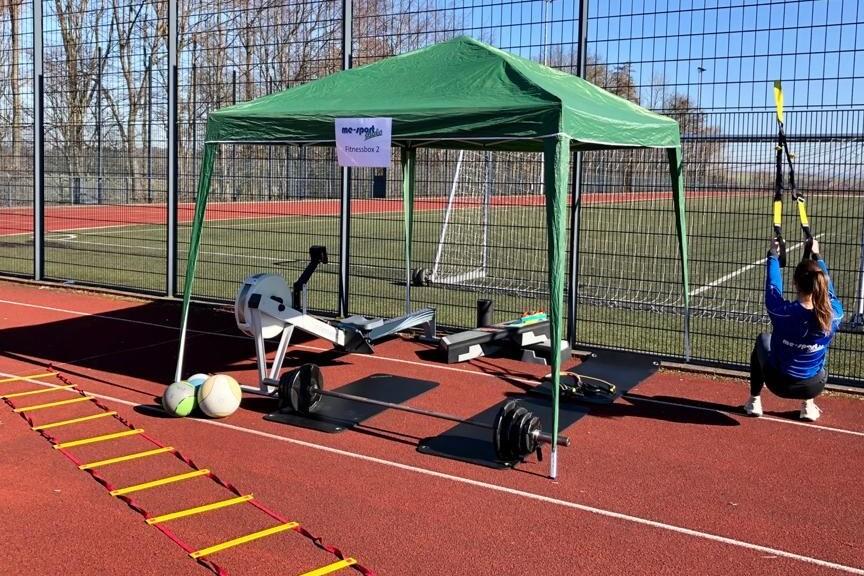 Sportbereich mit grünem Zelt, Fitnessgeräten, einer Person mit TRX-Bändern und Trainingsmaterial auf rotem Platz.