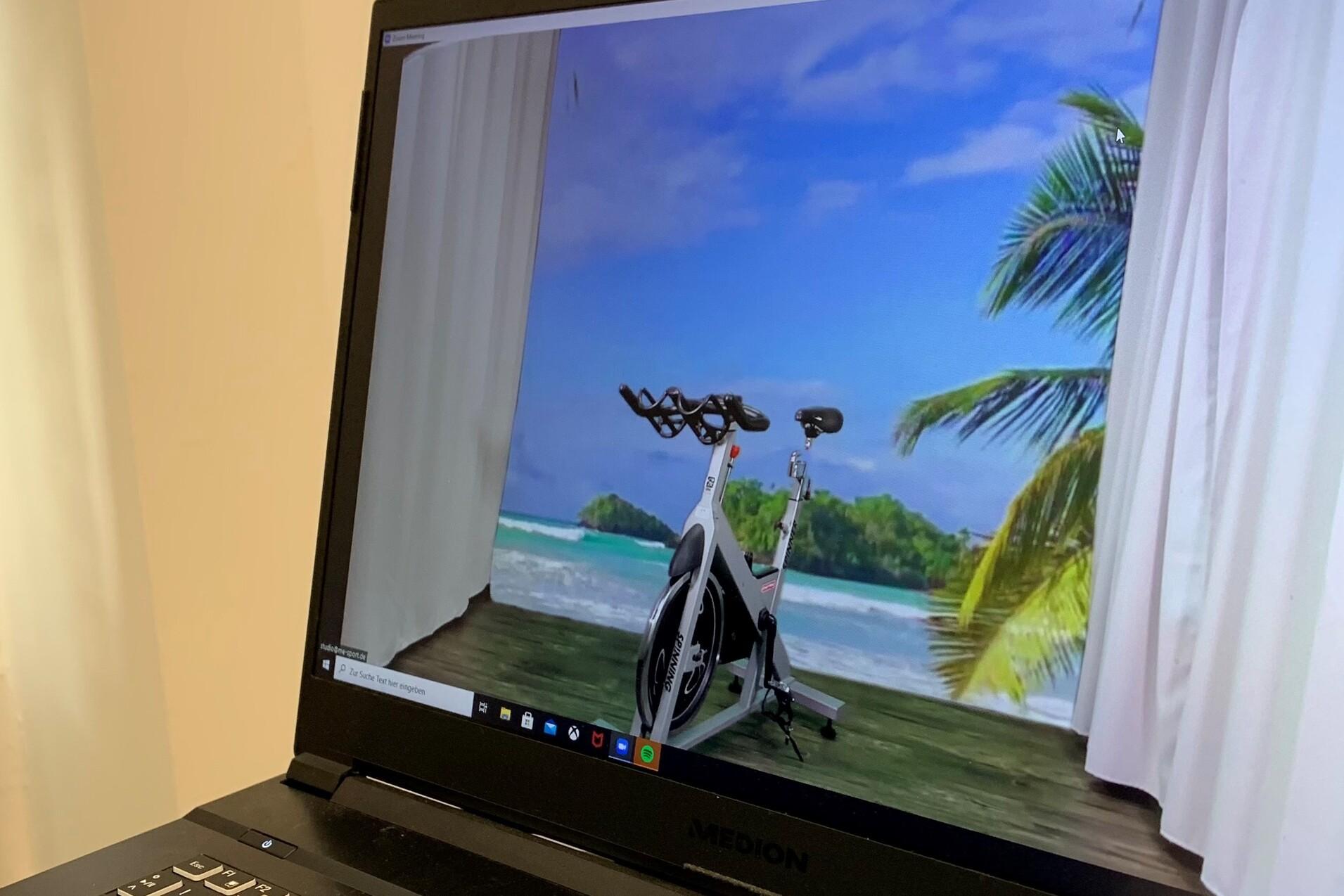 Virtuelles Fitnessgerät steht auf einem Tisch, im Hintergrund eine tropische Strandlandschaft mit Palmen.