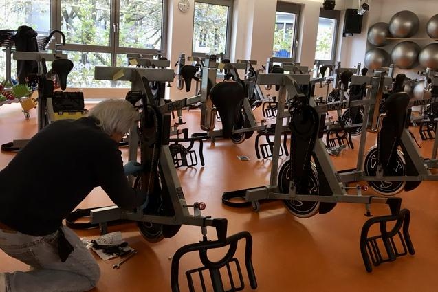 Mann schraubt an einem Fitnessfahrrad in einem hellen Raum mit mehreren &auml;hnlichen Fahrr&auml;dern und gro&szlig;en Fitnessb&auml;llen.