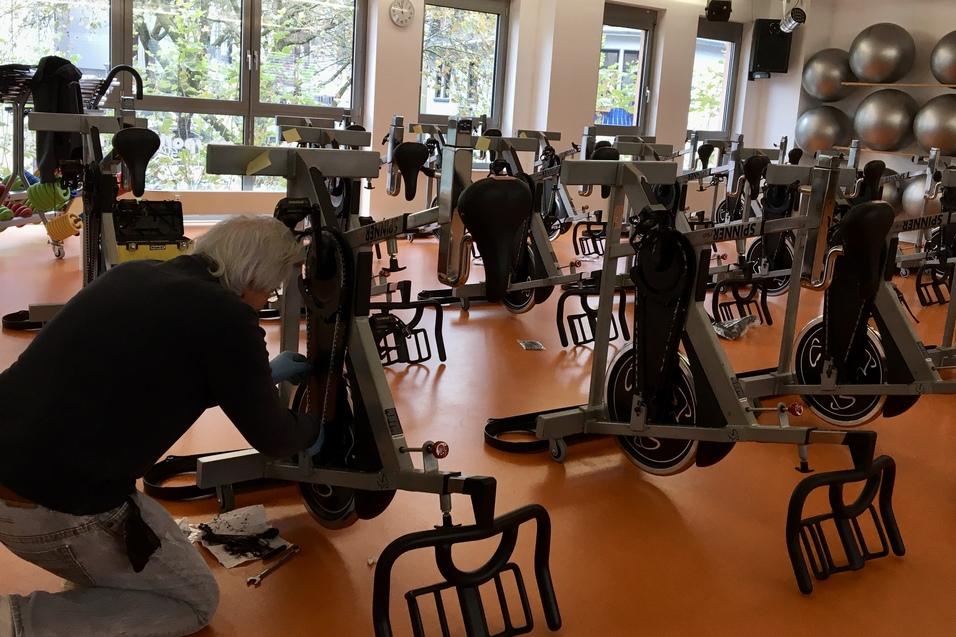 Mann schraubt an einem Fitnessfahrrad in einem hellen Raum mit mehreren ähnlichen Fahrrädern und großen Fitnessbällen.