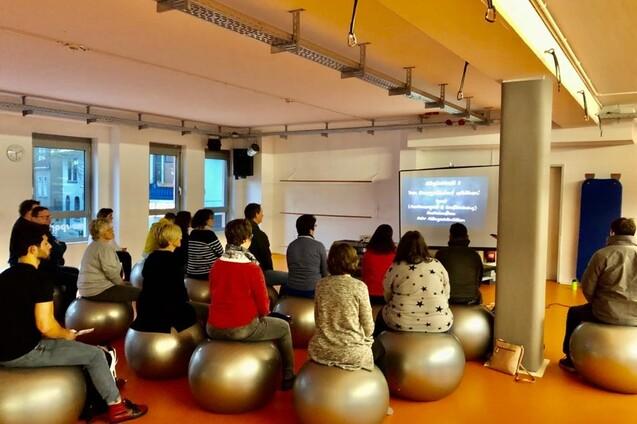 Gruppe von Menschen auf Gymnastikbällen sitzt und blickt auf einen Bildschirm mit Text in einem hellen Raum.