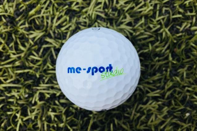 Weißer Golfball mit blauer und grüner Beschriftung "me-sport studio" auf grünem Kunstrasen.