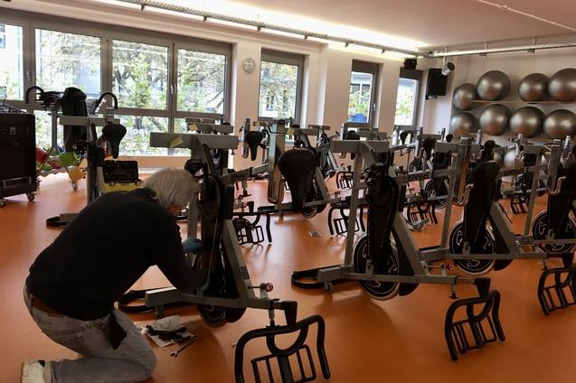 Mann schraubt an einem Fitnessfahrrad in einem hellen Raum mit mehreren ähnlichen Fahrrädern und großen Fitnessbällen.