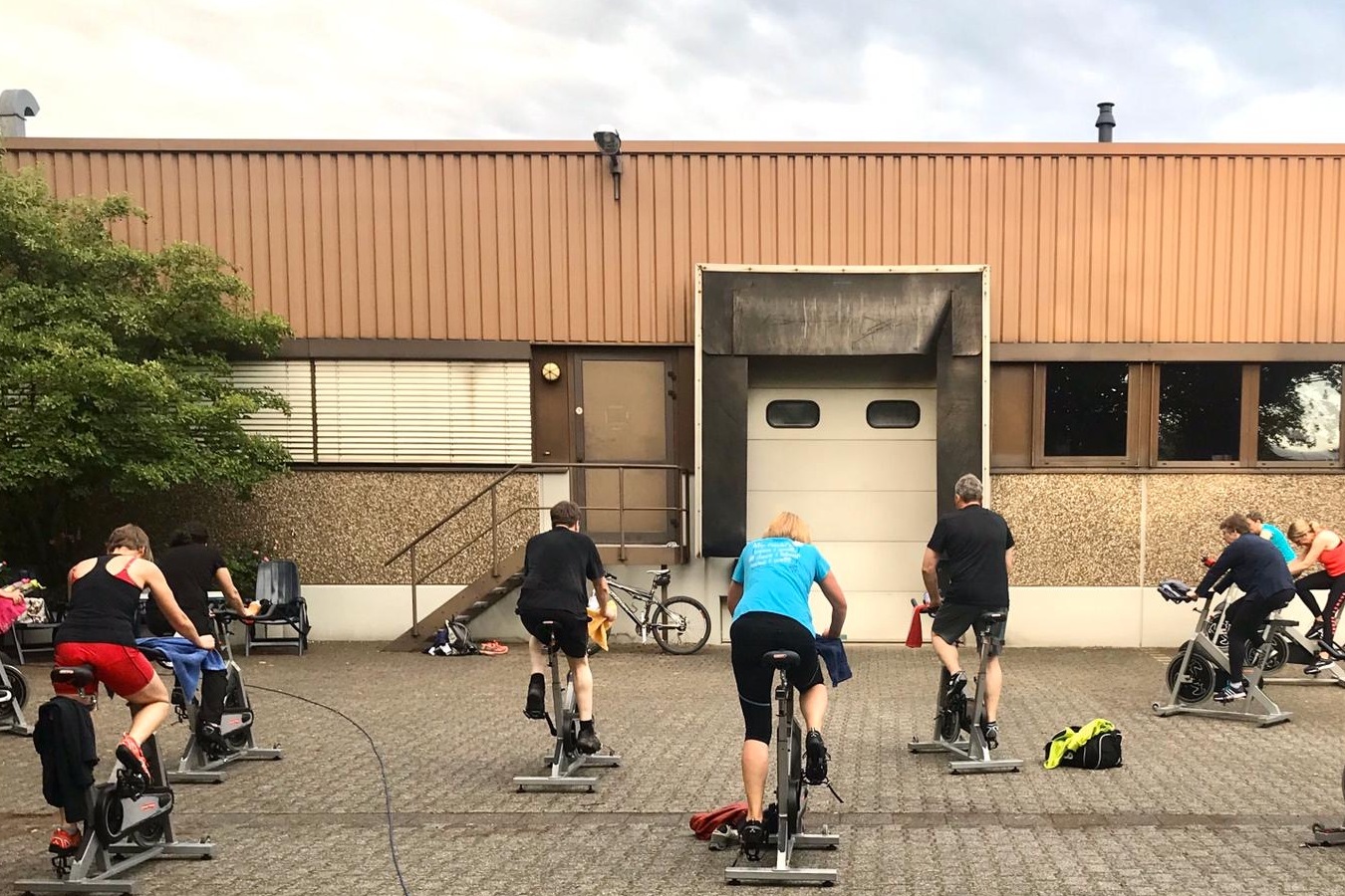 Gruppe von Personen beim Spinning unter freiem Himmel vor einem Geb&auml;ude, mit Fitnessbikes auf einem gepflasterten Boden.