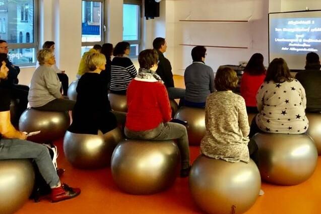 Gruppe von Menschen auf Gymnastikb&auml;llen sitzt und blickt auf einen Bildschirm mit Text in einem hellen Raum.