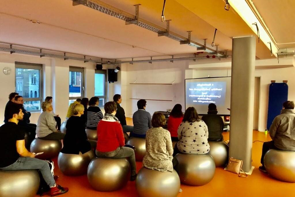 Gruppe von Menschen auf Gymnastikbällen sitzt und blickt auf einen Bildschirm mit Text in einem hellen Raum.