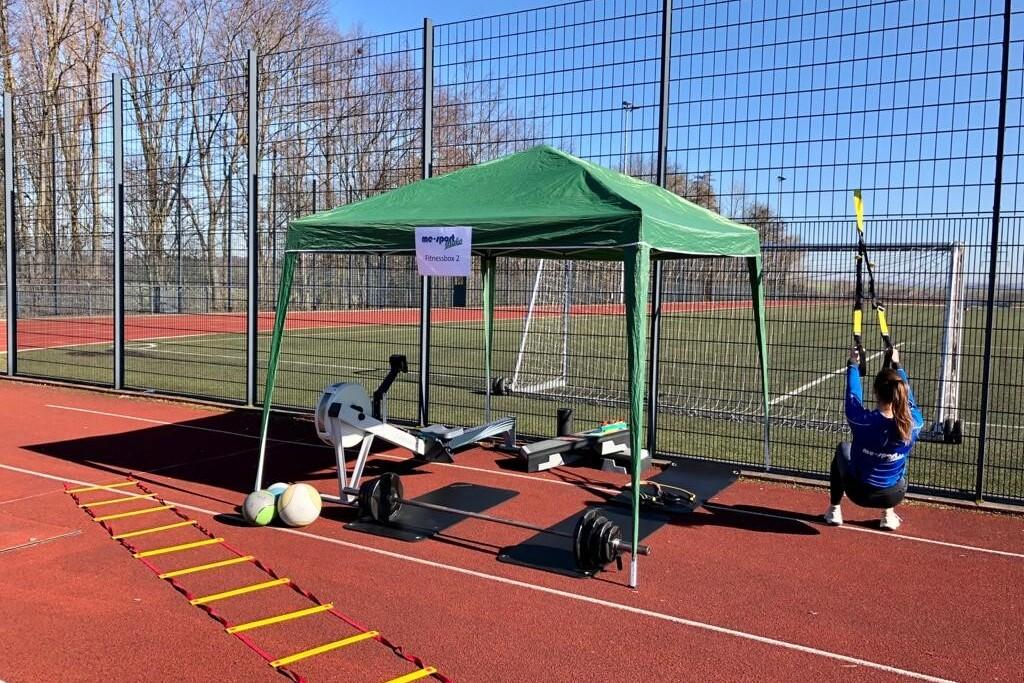 Sportbereich mit grünem Zelt, Fitnessgeräten, einer Person mit TRX-Bändern und Trainingsmaterial auf rotem Platz.
