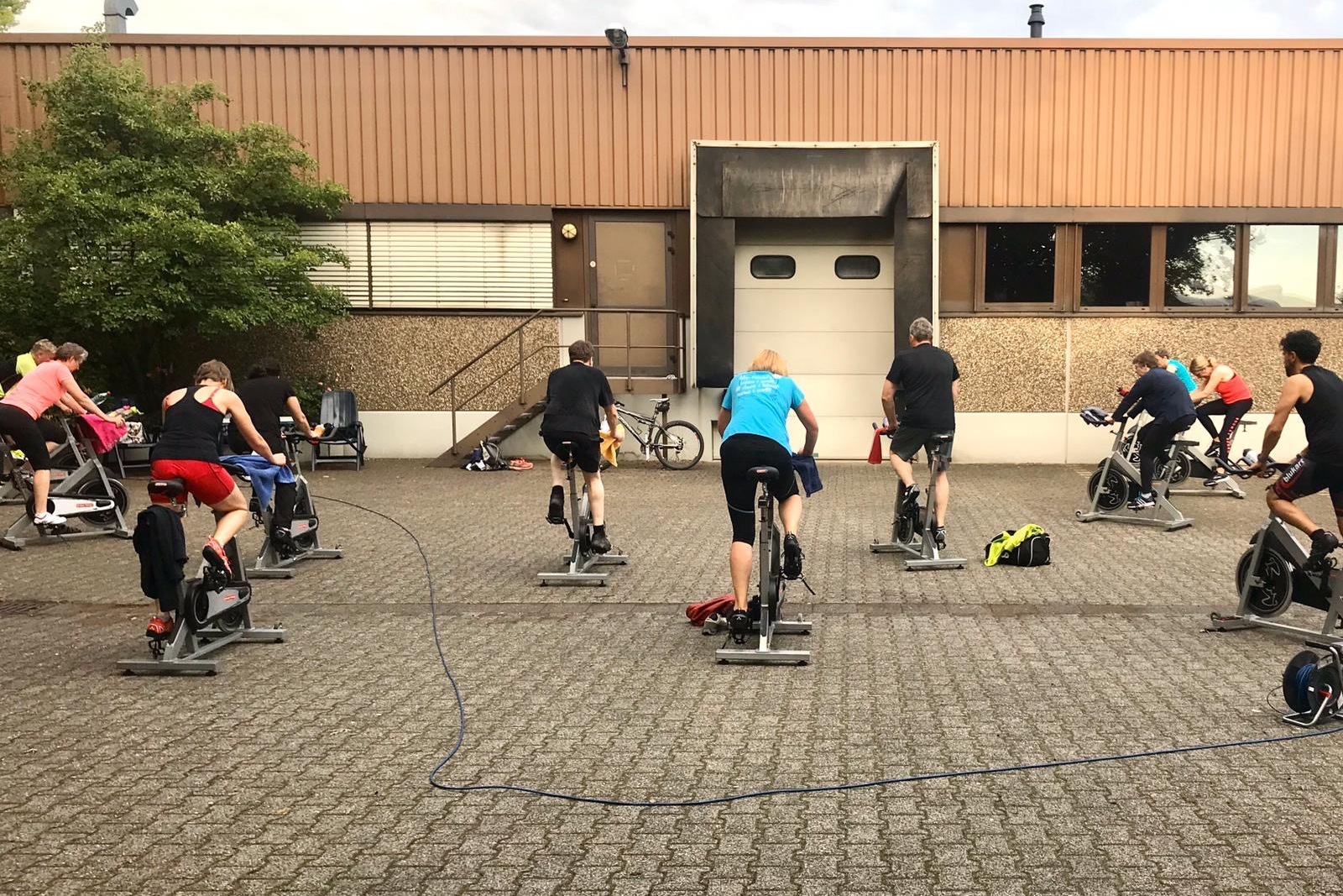 Gruppe von Personen beim Spinning unter freiem Himmel vor einem Gebäude, mit Fitnessbikes auf einem gepflasterten Boden.