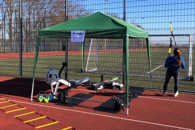 &Uuml;bungsecke auf einem Sportplatz mit einem gr&uuml;nen Zelt, Fitnessger&auml;ten und einer Person, die TRX-&Uuml;bungen macht.