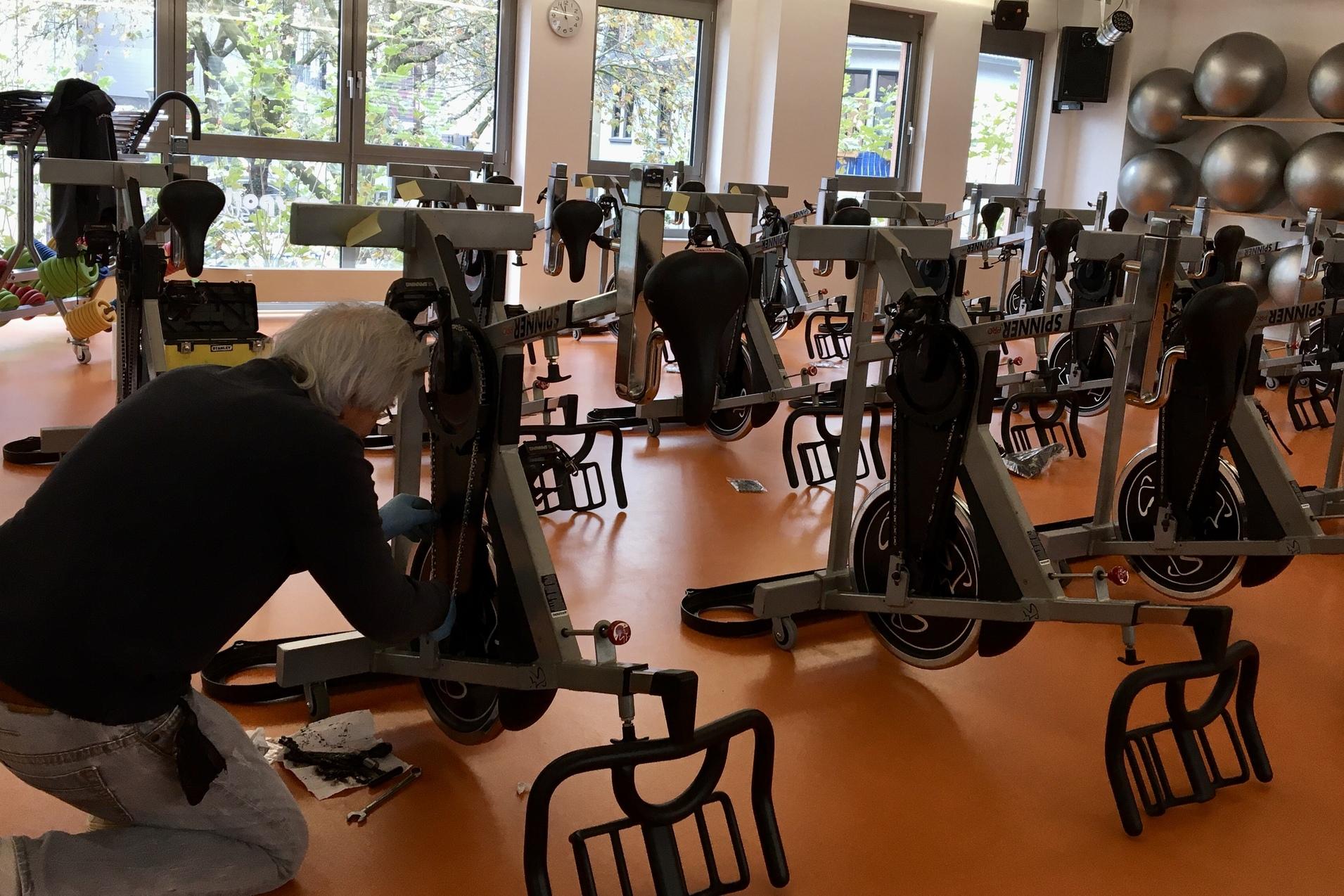 Mann schraubt an einem Fitnessfahrrad in einem hellen Raum mit mehreren &auml;hnlichen Fahrr&auml;dern und gro&szlig;en Fitnessb&auml;llen.