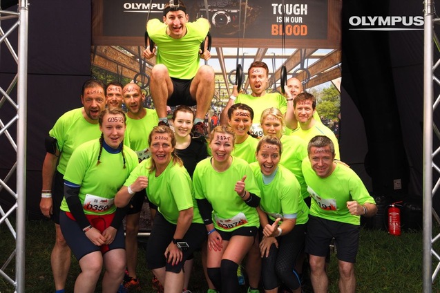 Gruppe von Teilnehmern in leuchtend grünen Shirts, zusammen lächelnd vor einem Banner mit dem Text „TOUGH IS IN OUR BLOOD“.