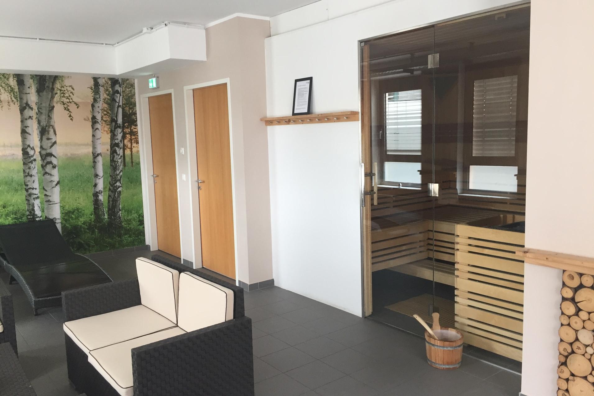 Moderne Wellness-Lounge mit Saunazugang, bequemen schwarz-weißen Loungemöbeln und einer Birkenwaldtapete.