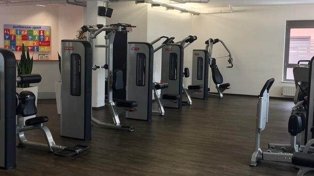 Fitnessraum mit mehreren Trainingsgeräten und großen Pflanzen in weißen Übertöpfen, helle Wände und Fenster im Hintergrund.