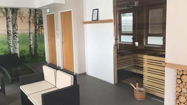 Moderne Wellness-Lounge mit Saunazugang, bequemen schwarz-weißen Loungemöbeln und einer Birkenwaldtapete.