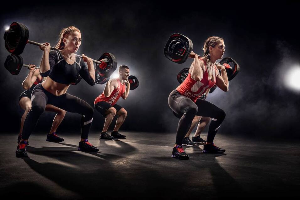Gruppentraining mit mehreren Personen, die Gewichtheben und Kniebeugen mit Langhanteln in einem beleuchteten Studio ausführen.