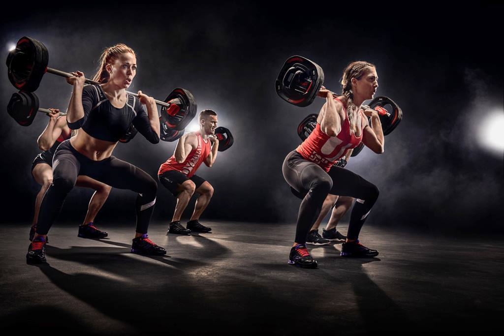 Gruppentraining mit mehreren Personen, die Gewichtheben und Kniebeugen mit Langhanteln in einem beleuchteten Studio ausführen.