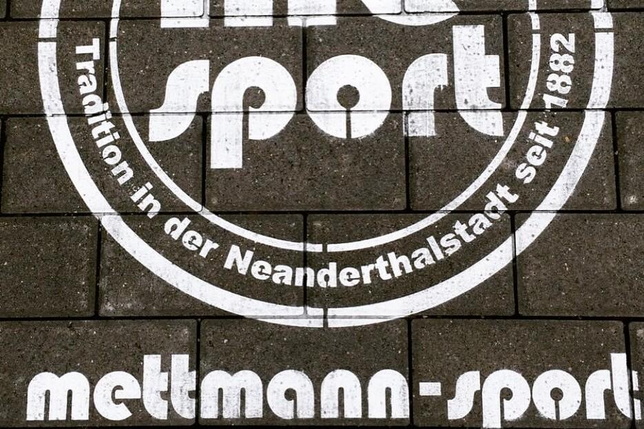 Weißes Logo mit der Aufschrift „me sport“ und dem Slogan „Tradition in der Neanderthalstadt seit 1882“ auf Pflastersteinen.