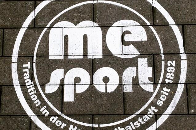 Wei&szlig;es Logo mit der Aufschrift &bdquo;me sport&ldquo; und dem Slogan &bdquo;Tradition in der Neanderthalstadt seit 1882&ldquo; auf Pflastersteinen.