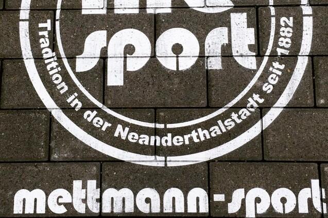 Wei&szlig;es Logo mit der Aufschrift &bdquo;me sport&ldquo; und dem Slogan &bdquo;Tradition in der Neanderthalstadt seit 1882&ldquo; auf Pflastersteinen.