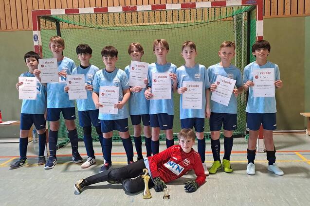 Gruppe von zehn Jugendlichen in Sportkleidung, die Urkunden und Pokale in einer Sporthalle stolz präsentieren.