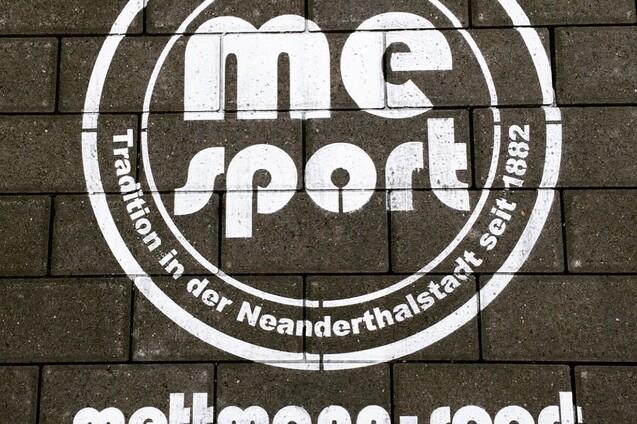 Weißes Logo mit der Aufschrift „me sport“ und dem Slogan „Tradition in der Neanderthalstadt seit 1882“ auf Pflastersteinen.