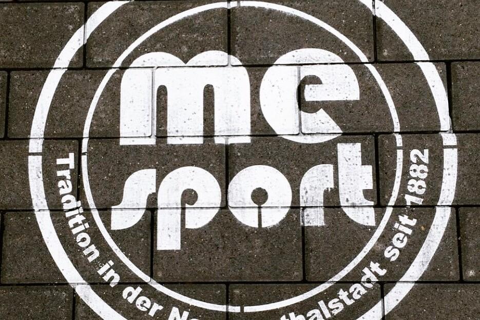 Wei&szlig;es Logo mit der Aufschrift &bdquo;me sport&ldquo; und dem Slogan &bdquo;Tradition in der Neanderthalstadt seit 1882&ldquo; auf Pflastersteinen.