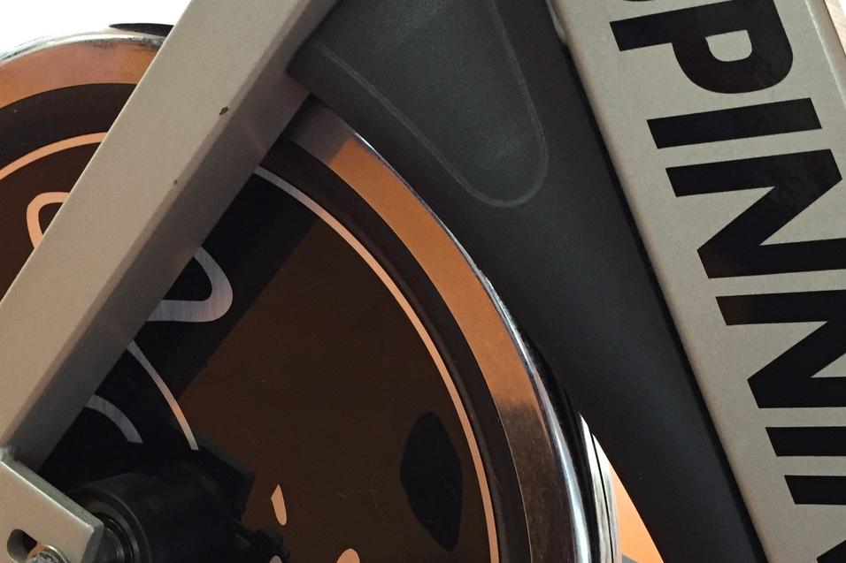 Detailansicht eines Spinning-Bikes mit dem Logo auf dem Schwungrad, vor einem orangefarbenen Untergrund.