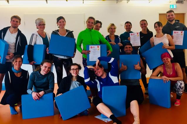 Gruppe von sportlich gekleideten Personen h&auml;lt blaue Platten und Urkunden in einem Raum mit orangefarbenem Boden.