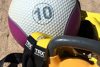 Fitnessger&auml;t auf Sandboden: Ein lilafarbener Medizinball, ein TRX-Gurt und eine orangefarbene Kettlebell.