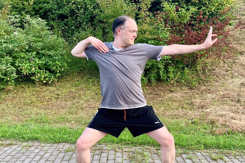 Mann in Sportkleidung steht in einer dynamischen Pose auf einem gepflasterten Weg, umgeben von Gr&uuml;nfl&auml;chen.