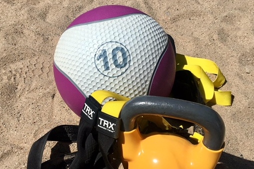 Fitnessgerät auf Sandboden: Ein lilafarbener Medizinball, ein TRX-Gurt und eine orangefarbene Kettlebell.