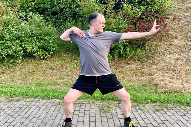 Mann in Sportkleidung steht in einer dynamischen Pose auf einem gepflasterten Weg, umgeben von Gr&uuml;nfl&auml;chen.