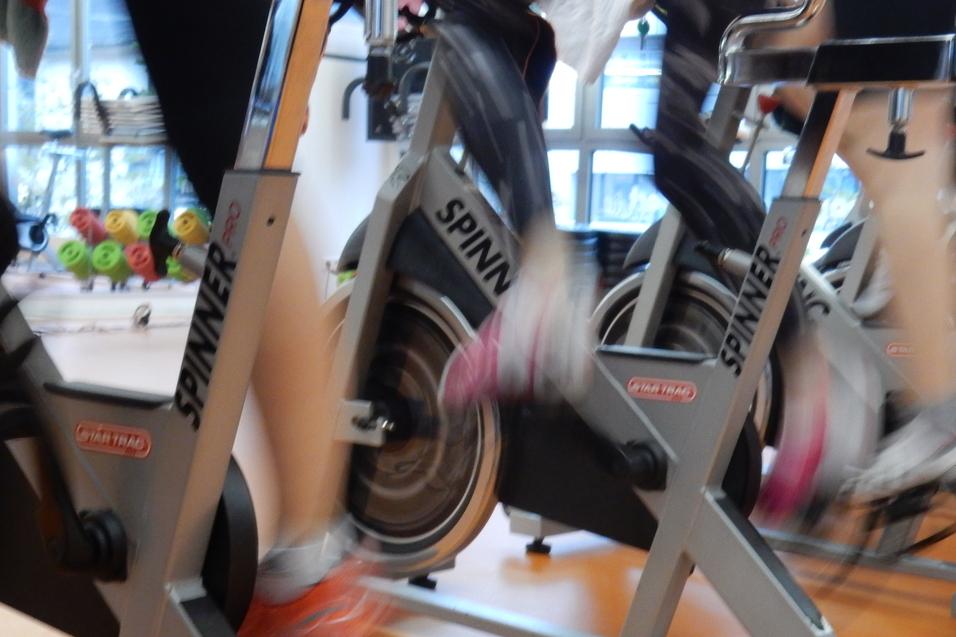 Bewegte Beine von Personen beim Spinning-Training auf stationären Fahrrädern in einem Fitnessstudio.