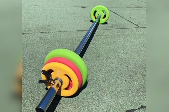 Langhantel mit bunten Gewichten auf einem grünen Sportboden, seitlich liegend und im Sonnenlicht.