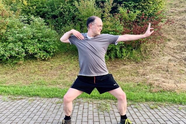 Mann in Sportkleidung steht in einer dynamischen Pose auf einem gepflasterten Weg, umgeben von Grünflächen.