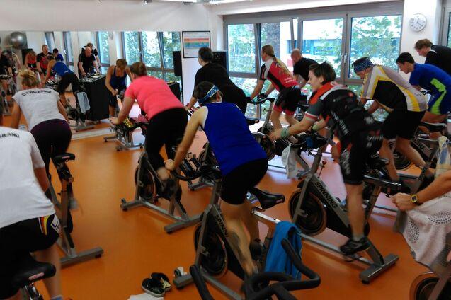 Gruppe von Menschen beim Indoor-Cycling in einem Fitnessstudio mit mehreren Spinning-Bikes und großen Fenstern.