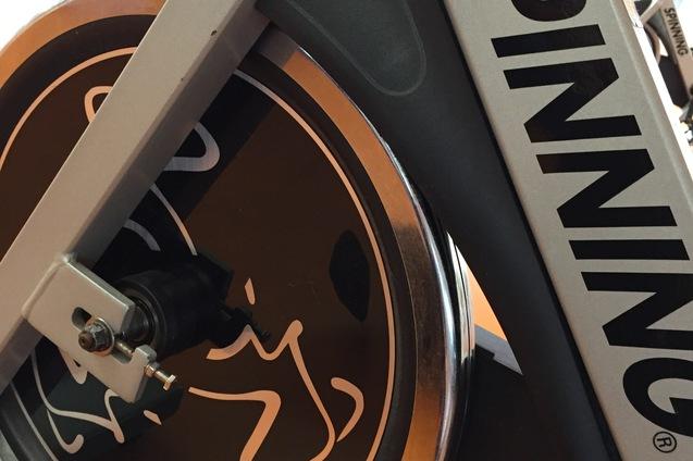 Detailansicht eines Spinning-Bikes mit dem Logo auf dem Schwungrad, vor einem orangefarbenen Untergrund.